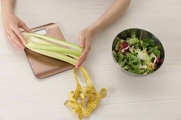 Woman weighting celery on kitchen scale at white wooden table, top view