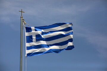 The Greek national flag flies against a blue sky, hung on a metal pole with a cross on top