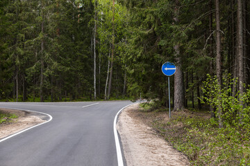 An empty asphalt road winding through a green forest, traffic sign directing left