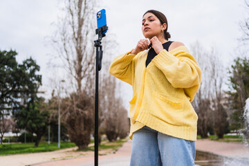 Latin influencer adjusting cardigan while filming content for social media with smartphone on tripod in a park