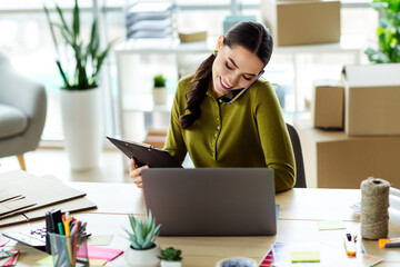 Busy businesswoman managing online orders while working in her home office surrounded by packing...