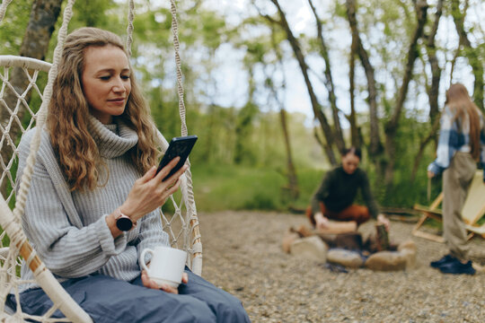 Middle-aged woman enjoying healthy lifestyle outdoors, sitting on swing with coffee, using phone in nature surrounded by trees, reflecting wellness and active wellbeing.