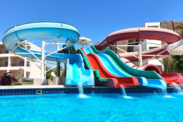 Bright and colorful water slides splash into a crystal-clear blue pool on a sunny day at a resort,...