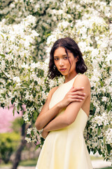 Attractive young woman in a yellow dress posing gracefully near blooming white flowers in a vibrant...