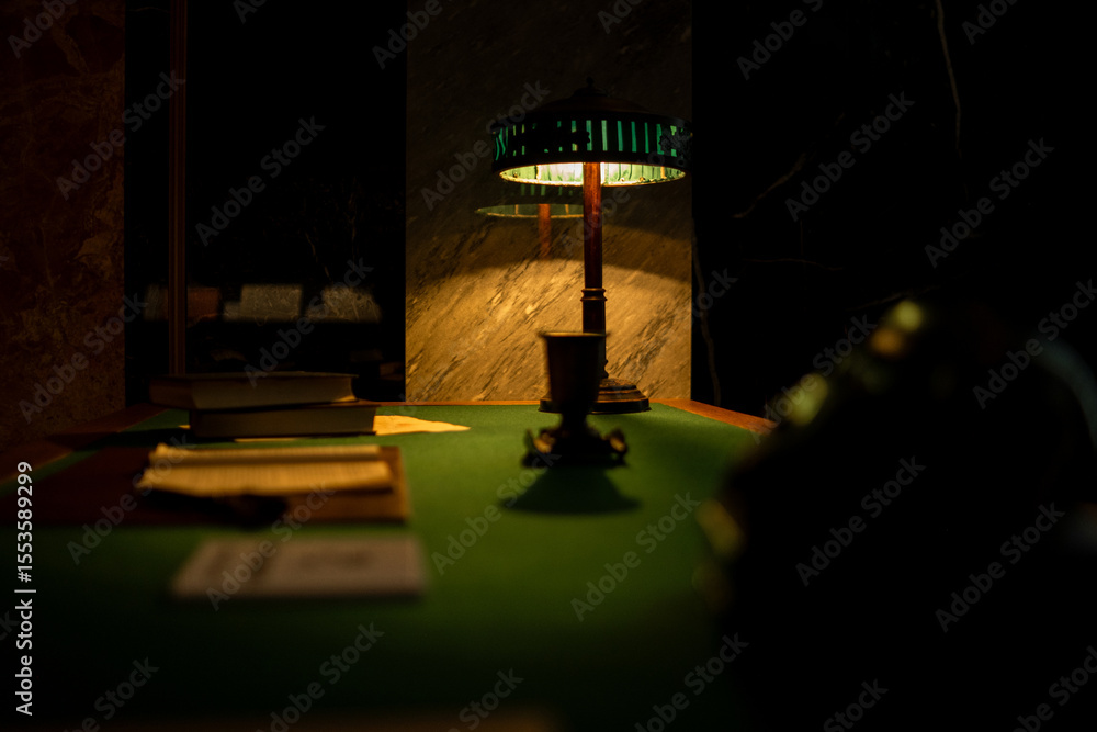 Poster warm glow from a vintage lamp illuminates a dark table set with books and documents in a quiet study