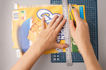 Hands cutting cereal box with utility knife and metal ruler on cutting mat. DIY craft material preparation using recycled cardboard. Step of creative handmade project. Top view composition,