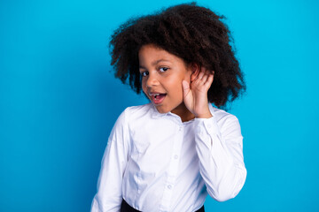 Curious young girl holding her hand to her ear, dressed in a white shirt, standing against a...