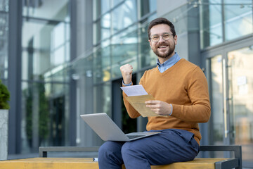 Portrait of a happy young man sitting on a bench outside, holding a laptop on his lap, an open...