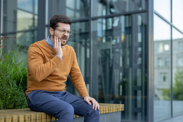 An office worker sits on a bench outside an office building and holds his cheek with his hand, feeling a severe toothache