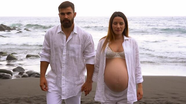 Happy young couple walking on the beach and kissing each other in front of camera - Love, pregnancy and parenthood concept  