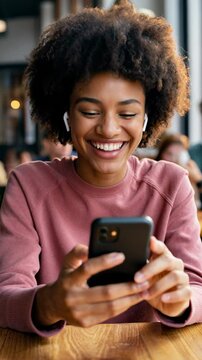 Smiling young woman in a pink sweater having a joyful video call with her best friend in a cozy caf&eacute;, holding her smartphone and wearing wireless earbuds &ndash; Generative AI