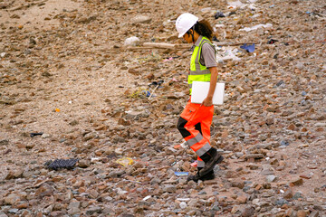 Female environmental engineer surveys rocky shoreline littered with waste, holding a laptop to...