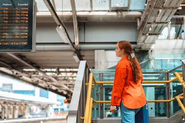 Woman waiting at train station for platform information in vibrant orange jacket during daytime, with digital schedule board in background