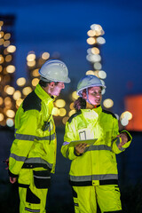 Two engineers in reflective safety gear review plans on a tablet during a night shift at an...