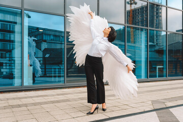 Woman poses elegantly with large white wings in a modern urban setting during an overcast day...