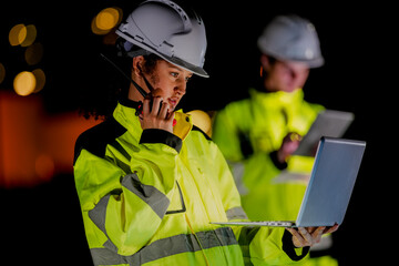 Female engineer in high visibility jacket uses a radio while analyzing data on a laptop during a...