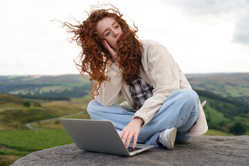 Young woman sits on a rocky outcrop in the countryside, using a laptop and gazing thoughtfully into...
