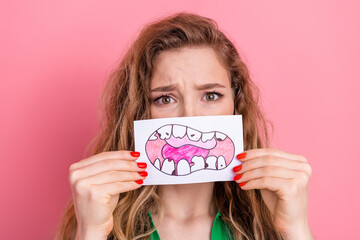 Young woman holding a drawing of teeth against a pink background, expressing worry and concern in a...