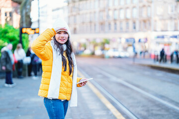 Young woman in yellow jacket stands at tram stop in city, looking around and checking her phone for...