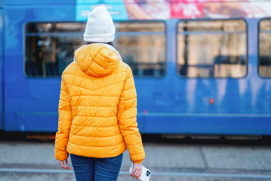 Woman wearing a yellow jacket waits at a tram stop while holding a smartphone in a vibrant city setting during daytime