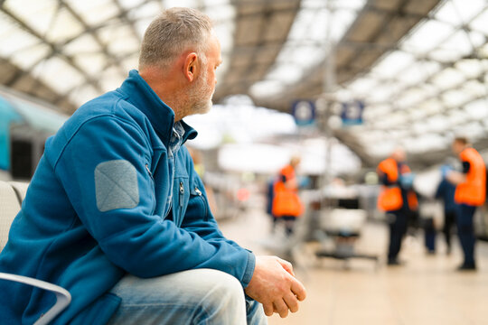 Man waiting in a busy train station during daytime, observing staff in bright uniforms assisting passengers nearby