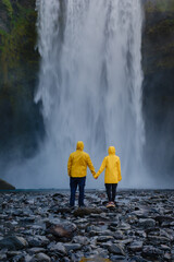 Couple holding hands in yellow raincoats by breathtaking Icelandic Skogafoss waterfall