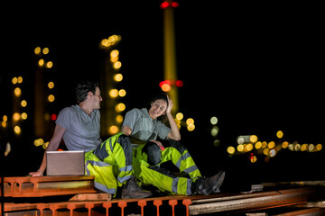 Two industrial workers take a break during the night shift at a construction site, sitting on steel...