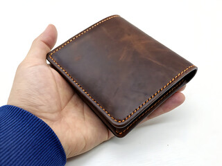 A hand holding a brown leather wallet with orange stitching on a white background in a studio shot