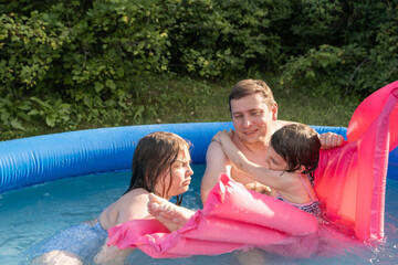 Children playing in inflatable pool on summer day. Two girls laughing, splashing water with father....