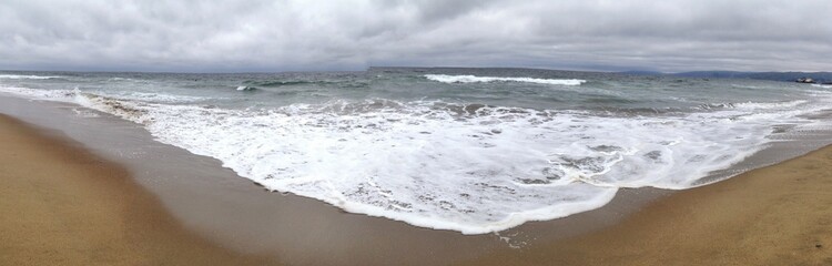Wide panorama of ocean waves reaching the sandy beach under cloudy skies