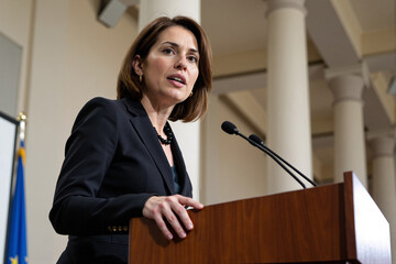 Powerful female politician, lawyer, or professor in a black blazer speaking with conviction at a podium in front of large columns in a government or university building