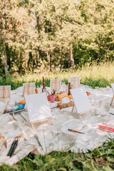 Closeup of picnic painting station with easels, blank canvases, and art supplies.	Detailed view of wooden mini-easels with white canvases, paintbrushes, cups with pencils, wine bottles and snack trays