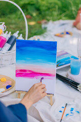 Brush glides across canvas creating pastel horizon tones during picnic painting workshop. Artist surrounded by grass, fabric, paint supplies and drinks. Summer creativity session with close-up view.
