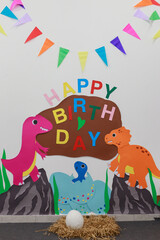 Obraz premium Dinosaur-themed birthday wall with colorful paper cutouts, festive letters HAPPY BIRTHDAY, cartoon pink orange dinosaurs, bright triangular flags decoration.