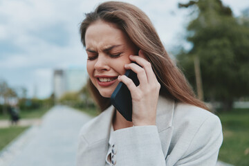 Young woman in beige jacket upset talking on phone outdoors, emotional distress, natural...