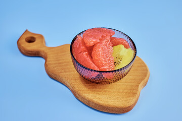 transparent vitamin bowl with grapefruit and kiwi pulp on a wooden pear-shaped board on a blue background close-up