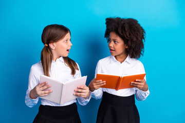 Two young schoolgirls with open book engaging in animated conversation on a bright blue background