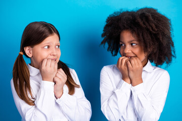 Two charming schoolgirls in white shirts nervously biting nails in front of a vibrant blue...
