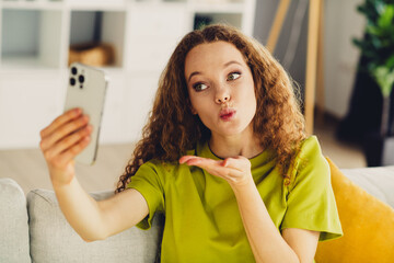 Young woman with curly hair sends a kiss to her phone camera while wearing a casual green shirt in...