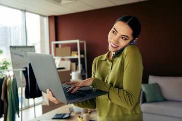 Young entrepreneur working in an atelier with a laptop multitasking phone call, planning and...