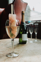 Close-up vertical shot of grapefruit popsicle champagne cocktail in wine glass with mint sprig, champagne bottle and flutes on rustic wooden table