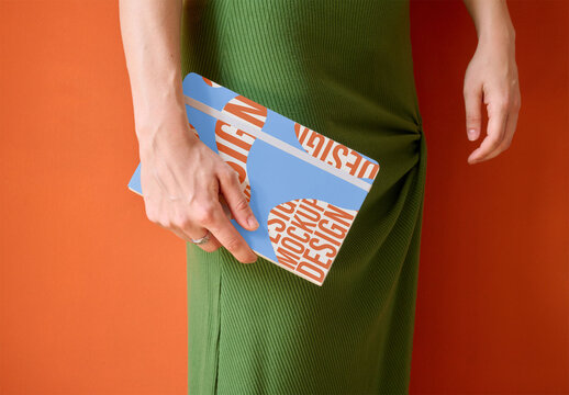 Womanholds a mockup of a notebook while wearing a green outfit against a warm orange backdrop, ready for creative ideas.