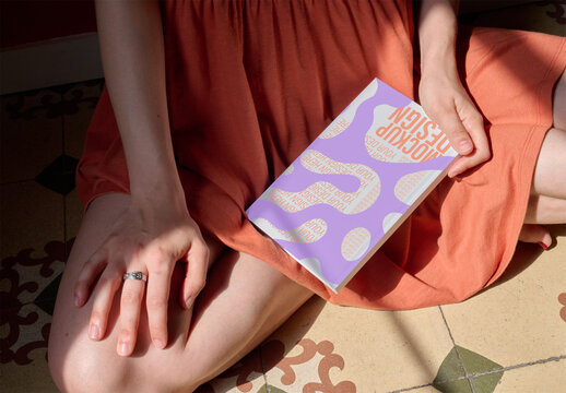 Woman sits on patterned tiles, holding a blank notebook in their lap, illuminated by natural light. PSD mockup.
