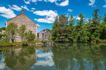 Historic 19th-century Canadian watermill with wooden buildings and stone structure, reflected in a calm river under a blue summer sky. Surrounded by forest and rural landscape.