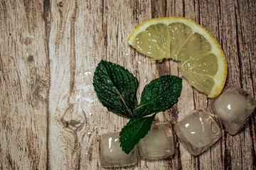 A fresh lemon slice, vibrant mint leaves, and glistening ice cubes arranged on a rustic wooden...