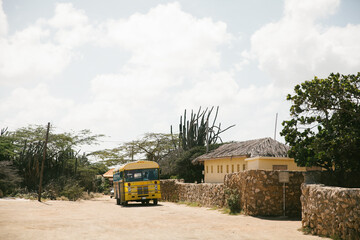 Bright yellow bus travels along dirt road near stone walls and greenery under cloudy sky in rural...