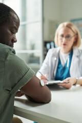 Obraz premium Middle aged Black man wearing glucose monitor on arm consulting with Caucasian female doctor in medical office, doctor holding clipboard and discussing diabetes management