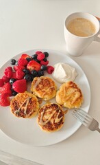 Homemade syrniki with berries and sour cream served on a white plate with a cup of coffee. Concept...