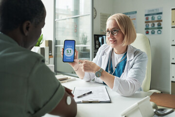 Middle aged Caucasian female doctor showing digital glucose meter to middle aged Black man during...