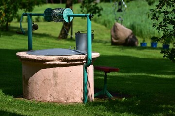 Old countryside water well with a green metal crank and a silver bucket. Surrounded by green grass...